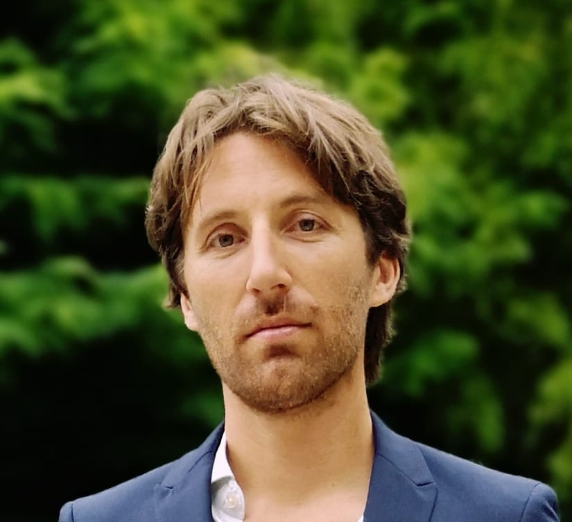 Photo of a man in his 30's, wearing a suit, with trees in the background.