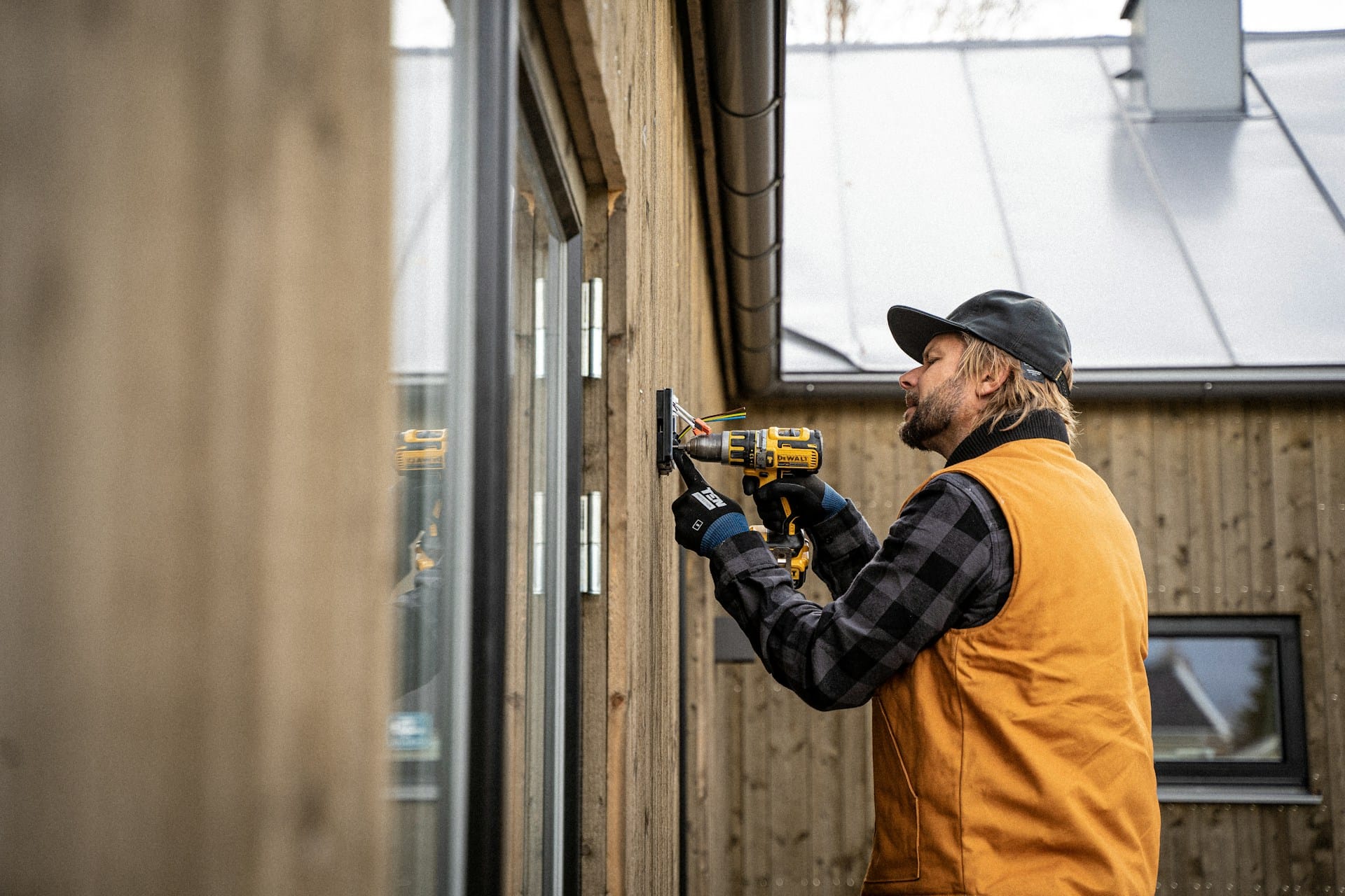 Un électricien avec gilet orange et casquette, en train de visser un boîtier électrique sur un mur extérieur de maison.