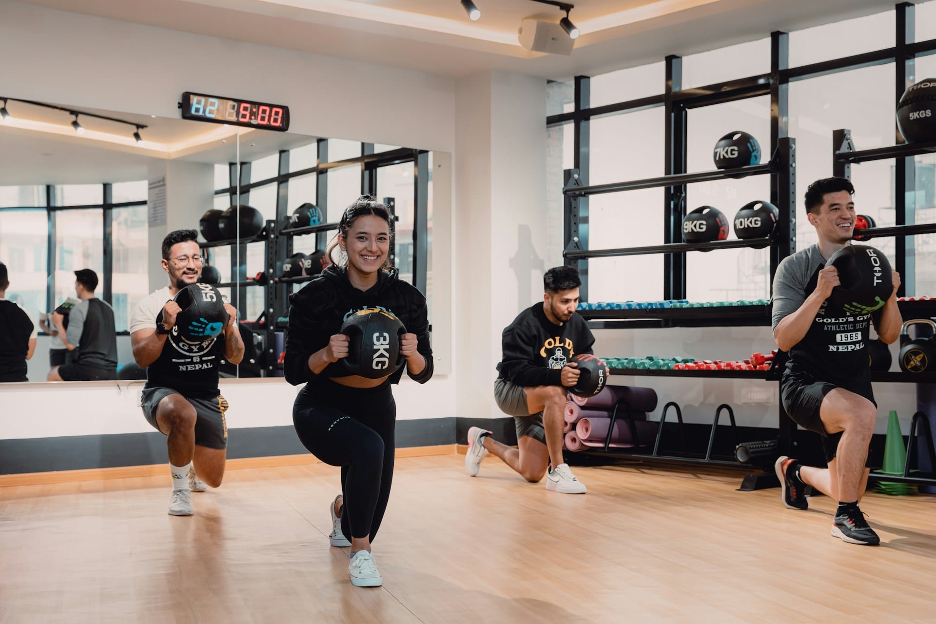 3 hommes et une femme lors d'une séance de sport dans un studio de fitness. Ils sont en position de fente genou fléchi, avec chacun un poids de plusieurs kgs dans les mains.