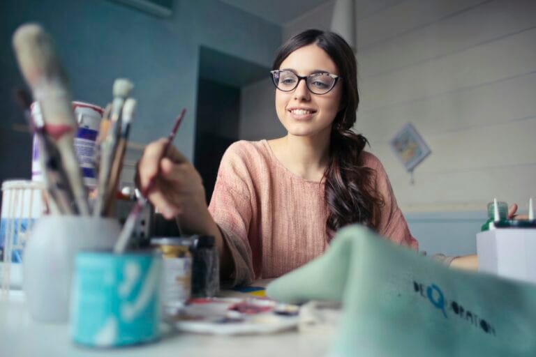 Une peintre avec le sourire en plein travail à sa table sur laquelle se trouvent plusieurs pots de peinture et des pinceaux.